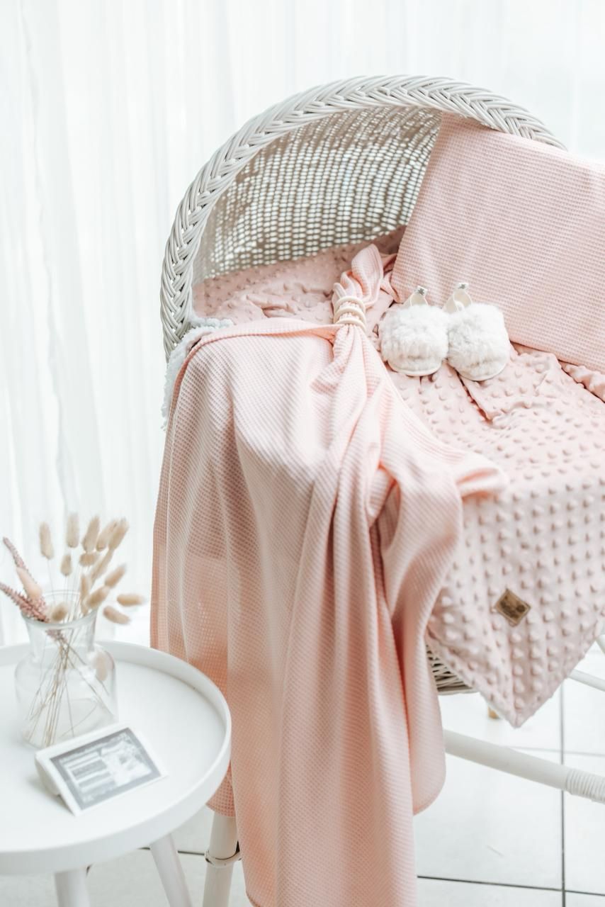 Soft pink baby blanket draped over white wicker bassinet with delicate bow detail in nursery setting.