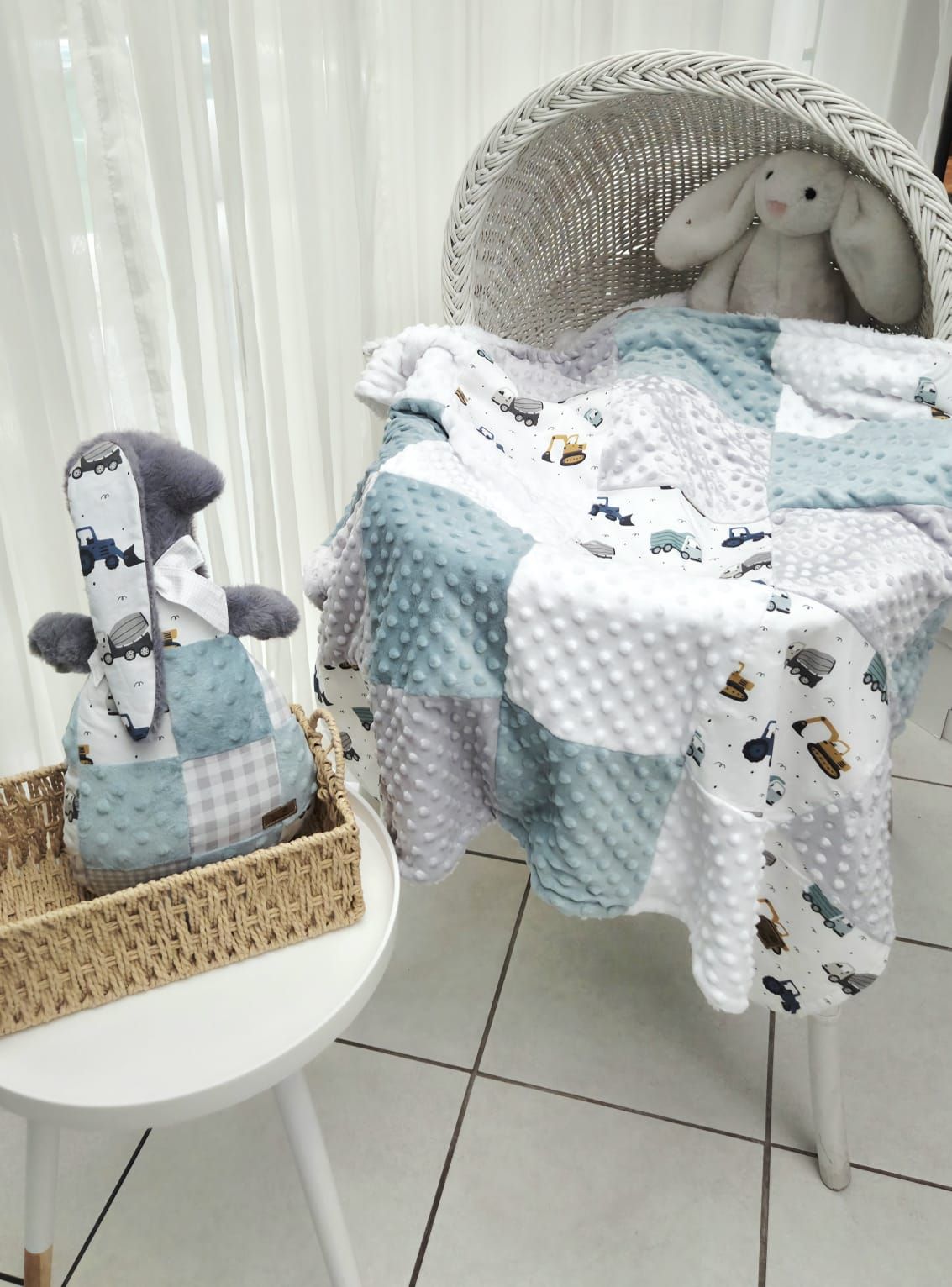 Teal and white patchwork baby blanket with vehicle print draped over wicker bassinet, with stuffed bunny and toy.