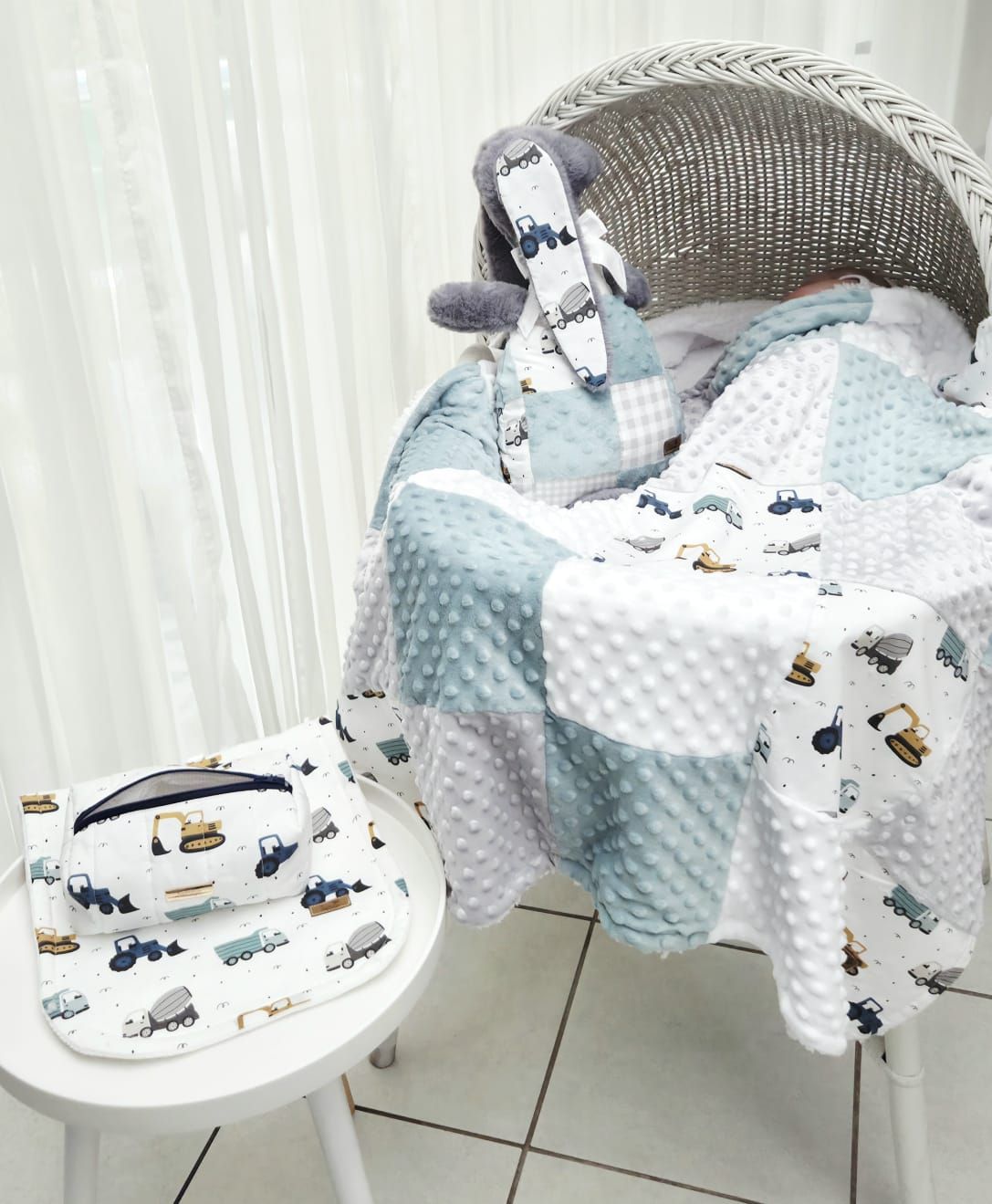 Baby boy patchwork blanket with construction vehicles in blue and white, displayed on a wicker chair with matching bib.