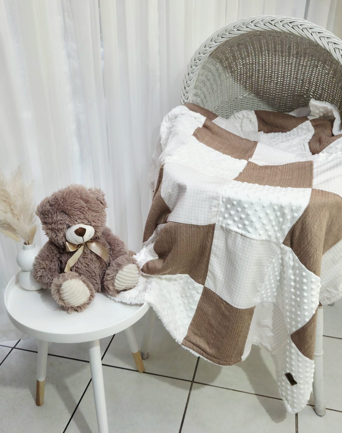 Brown and white checkered baby blanket draped over a wicker chair beside a teddy bear on a white stool.