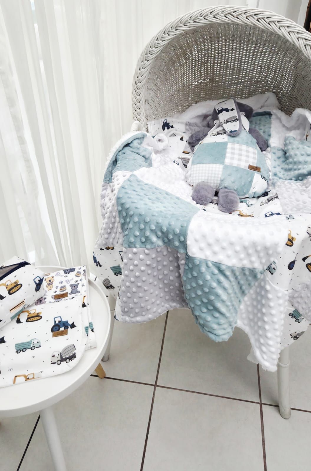 White wicker Moses basket with teal and white minky patchwork baby blanket, construction vehicle print bedding set.