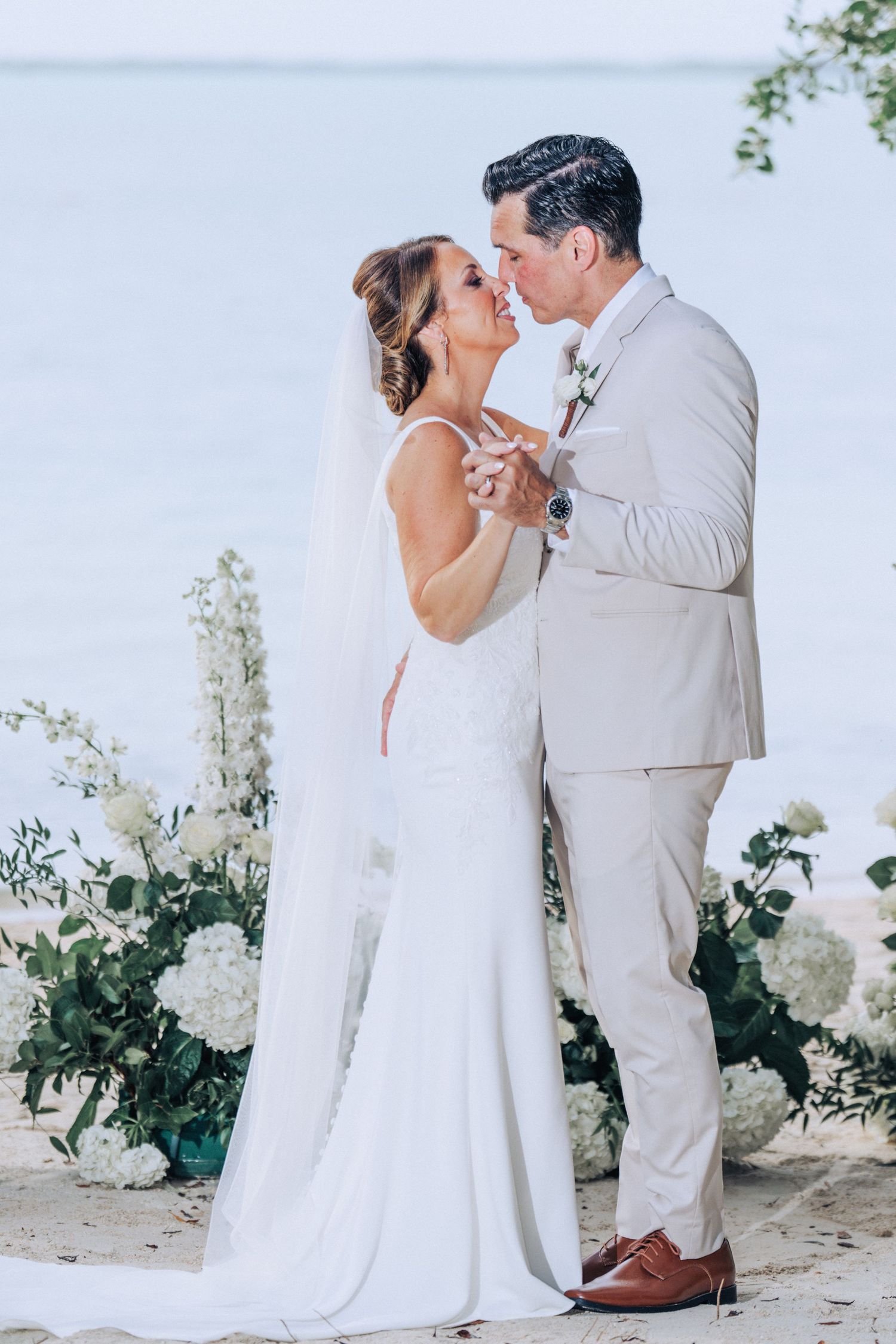 Romantic wedding couple in white attire share intimate moments during outdoor photo session with floral backdrop.