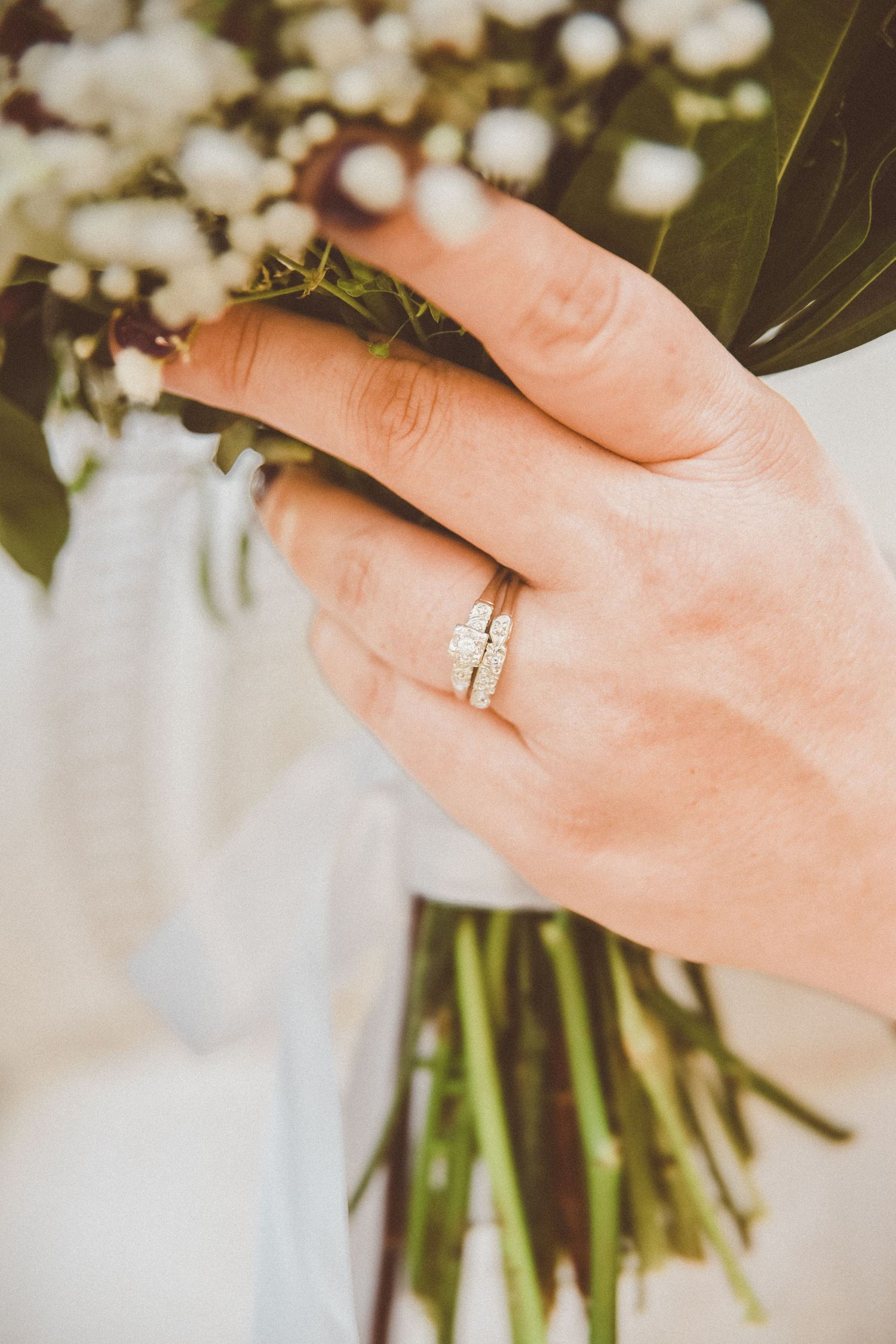 An image of a woman's hand wrapped around a bouquet of flowers, showing off both engagement and wedding rings.