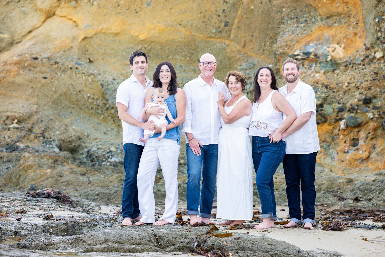 Dana Point photographer Extended family of six poses together on rocky coastal beach with golden cliffs in background.