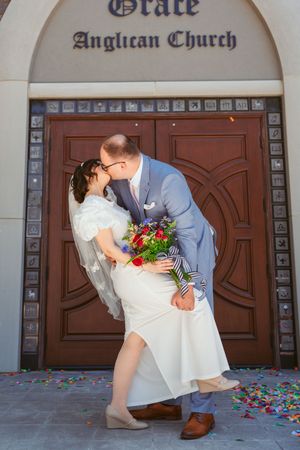 A romantic wedding couple shares a kiss in front of ornate wooden church doors at Grace Anglican Church with scattered flower petals.