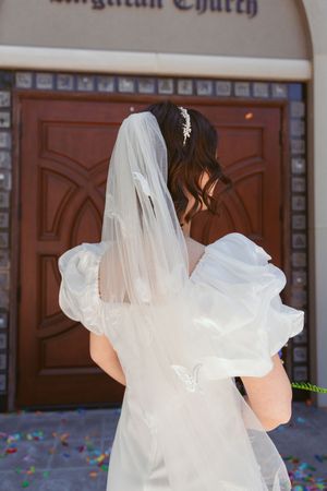 Back view of a bride in a flowing white veil and puffy dress sleeves standing before wooden church doors.