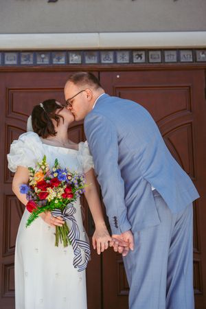 Newlyweds share a romantic kiss in front of wooden doors while bride holds vibrant multicolored wedding bouquet.