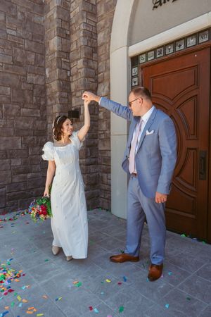 A couple in formal wedding attire share a joyful moment at a stone church entrance with colorful flower petals scattered on the ground.