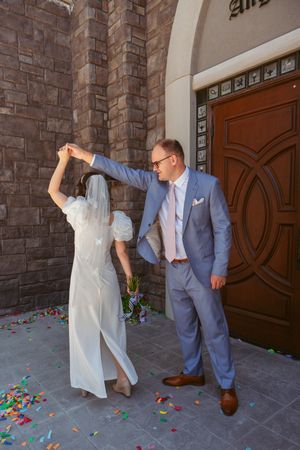 A groom in a gray suit twirls a bride in a white dress with veil under an arched doorway with scattered confetti.