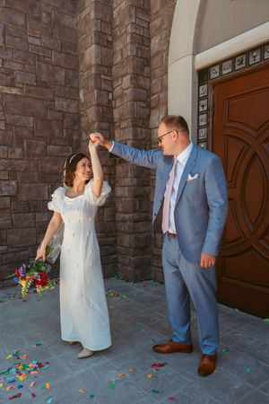 Wedding sequence showing couple playfully posing with confetti against stone church wall in blue suit and white dress.