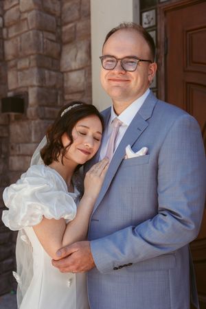 A romantic wedding portrait shows a couple embracing outside a stone building, with the bride in white and groom in gray suit.