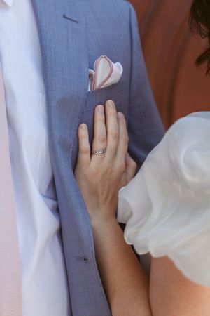 Close-up of a bride's hand with wedding ring resting on a light blue suit jacket with pink pocket square.