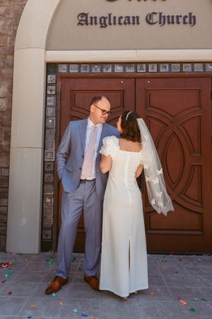 A couple in wedding attire share a moment in front of ornate wooden church doors with scattered rose petals.
