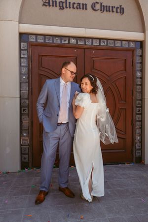 A couple taking playful wedding photos outside an Anglican church with brown wooden doors.