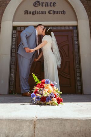 Vibrant bouquet of colorful flowers sits on concrete steps outside Grace Anglican Church with a couple in the background.