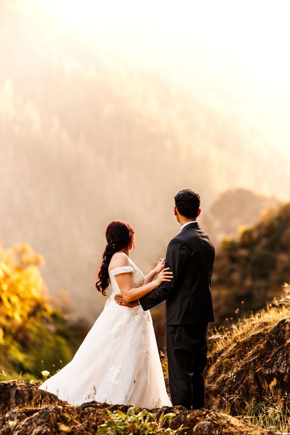 Bride and groom stand on a misty mountain cliff at golden hour, sharing a romantic moment in wedding attire.