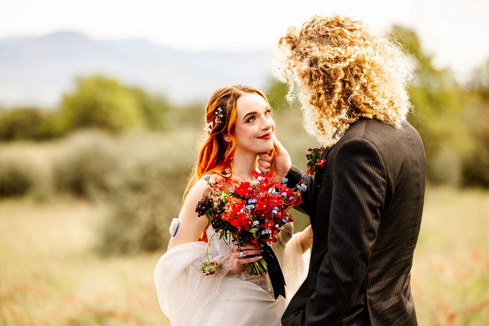 Two brides share a romantic moment in a field, one holding a vibrant red bouquet, mountains visible in the background.