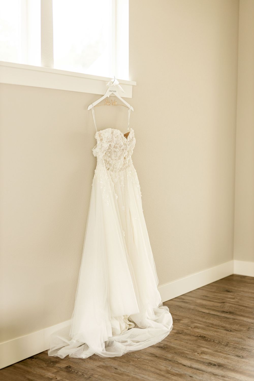 White wedding dress hangs elegantly on a white hanger against a neutral wall with rustic wooden flooring below.