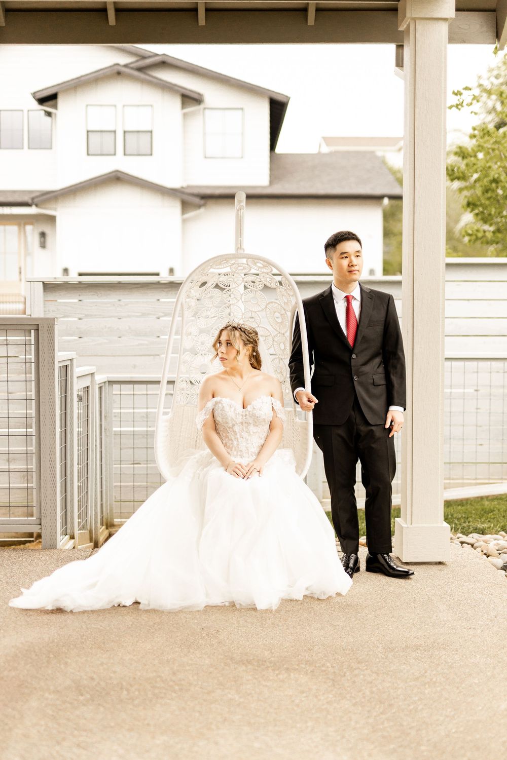 A couple stands together outdoors, with the bride sitting in a hanging chair wearing a white gown and the groom in a dark suit.