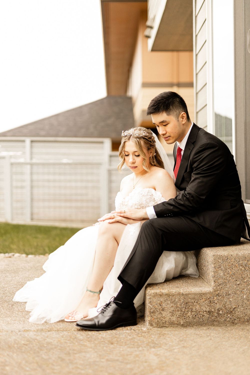 A bride and groom share an intimate moment sitting on concrete steps outside a building during their wedding day.
