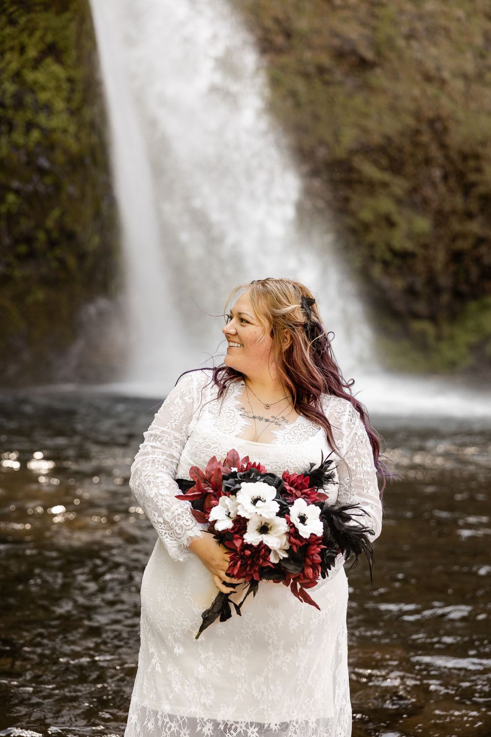 A bride in a white lace dress holds a red and white bouquet while standing in front of a majestic waterfall.