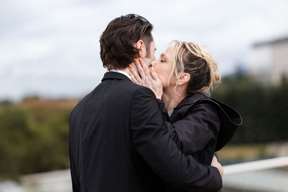 A romantic kiss between a couple on a rooftop, with the woman wearing black and the man in a dark suit.