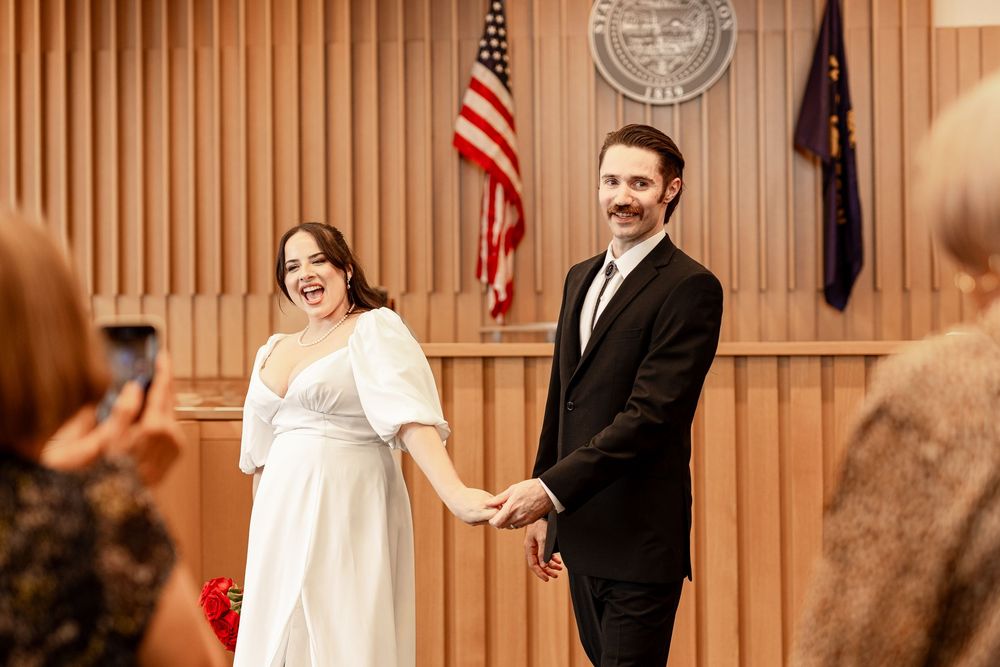 A joyful couple in a courthouse ceremony with American flag and state seal, bride in white dress and groom in black suit.