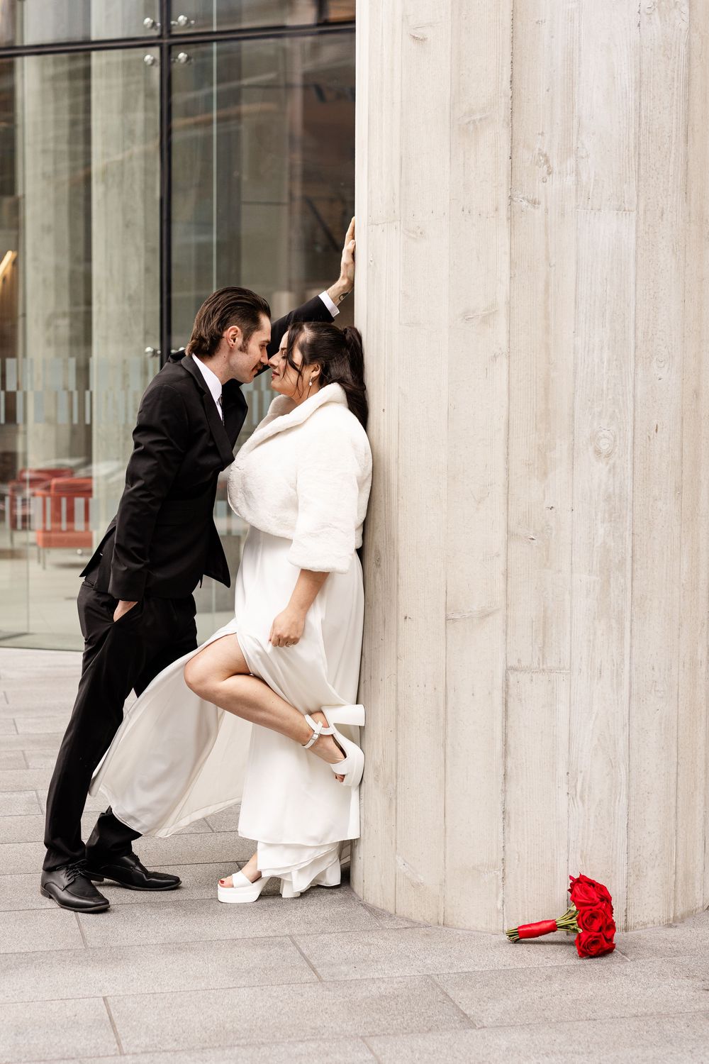 A romantic couple in wedding attire share an intimate moment against a stone wall with a red bouquet on the ground.