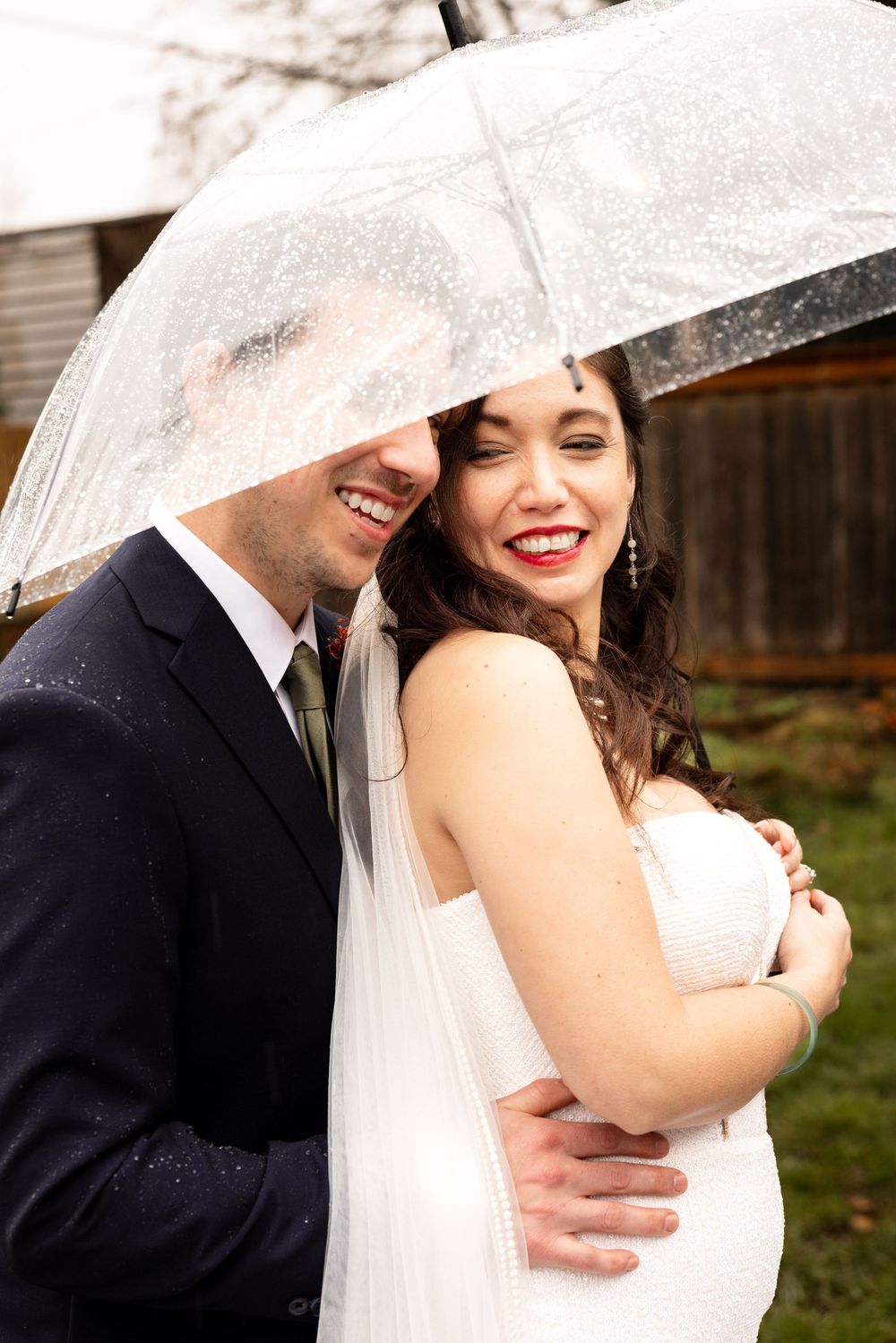 Bride and groom smiling under a clear umbrella in the rain, bride in white gown with red lips.