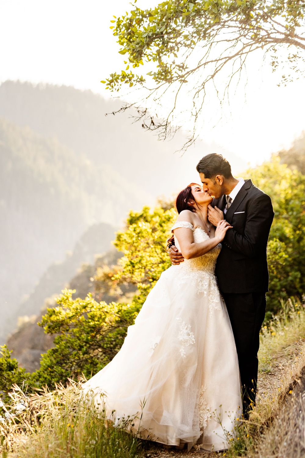 Bride and groom share a romantic kiss in a white lace gown and black suit with mountain scenery backdrop.