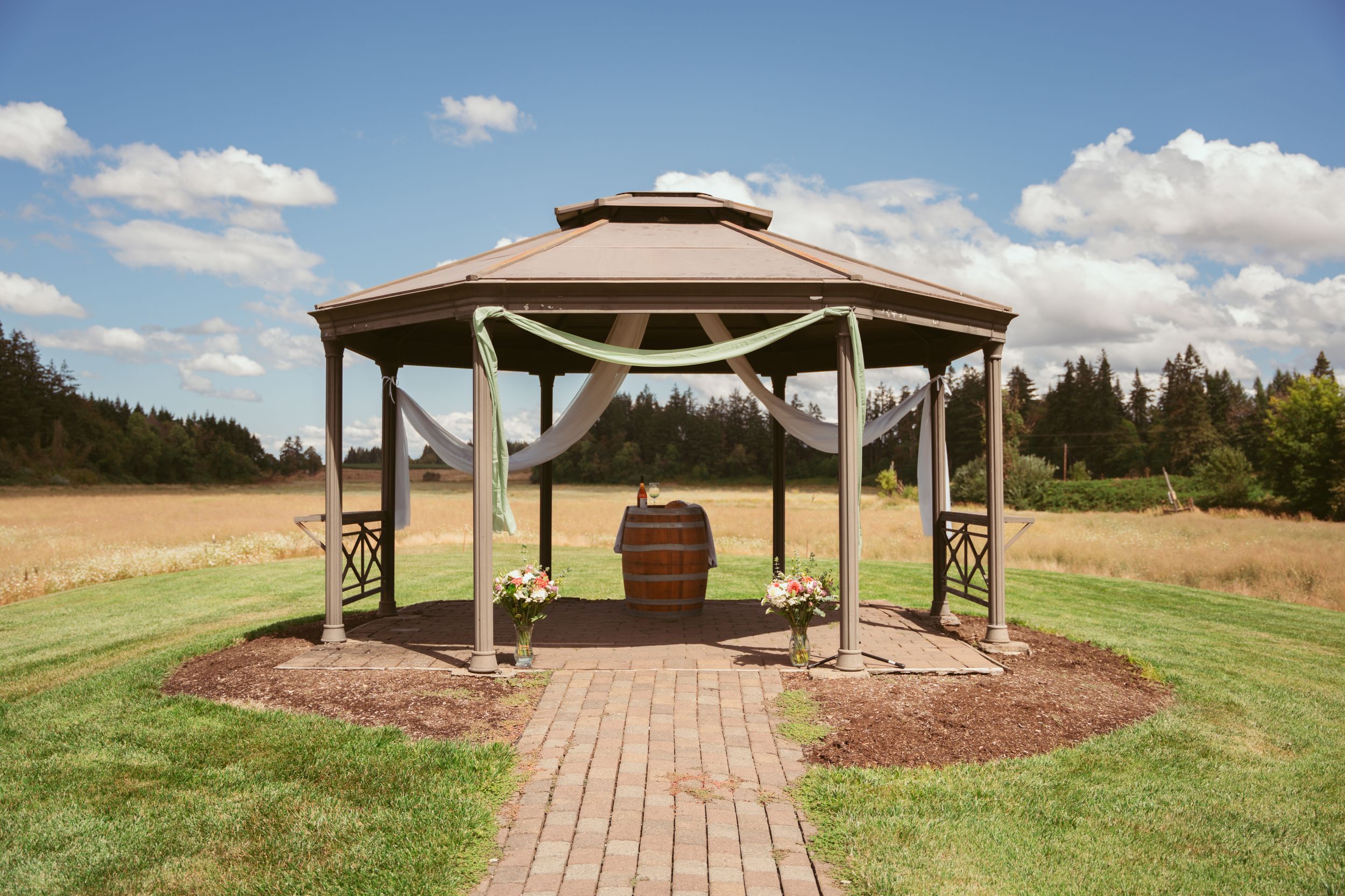 A wooden walkway leads to a rustic covered outdoor pavilion on a grassy hilltop with scenic mountain views in the distance.