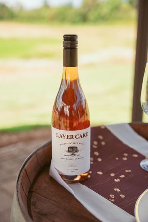 A bottle of Layer Cake wine sits on a rustic wooden barrel table with blurred outdoor background.