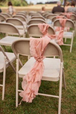 White folding chairs decorated with flowing peach-colored fabric ribbons arranged on grass for an outdoor wedding ceremony.