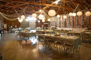 String lights and paper lanterns illuminate a rustic barn reception venue with wooden tables and chairs set for a wedding.
