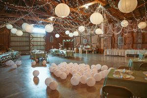 A rustic barn wedding venue decorated with white paper lanterns, string lights, and balloon arrangements on the floor.