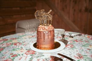 Gold drip wedding cake with topper on floral tablecloth featuring chocolate frosting and metallic accents.
