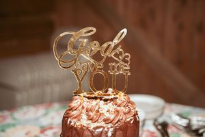 Elegant gold 'God Loves You' cake topper decoration on pink frosted dessert with blurred floral background.