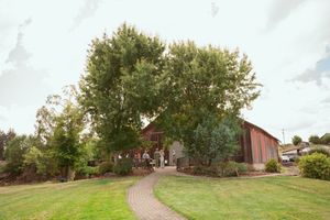 A rustic brown barn sits at the end of a curved brick pathway surrounded by lush trees and green lawn.