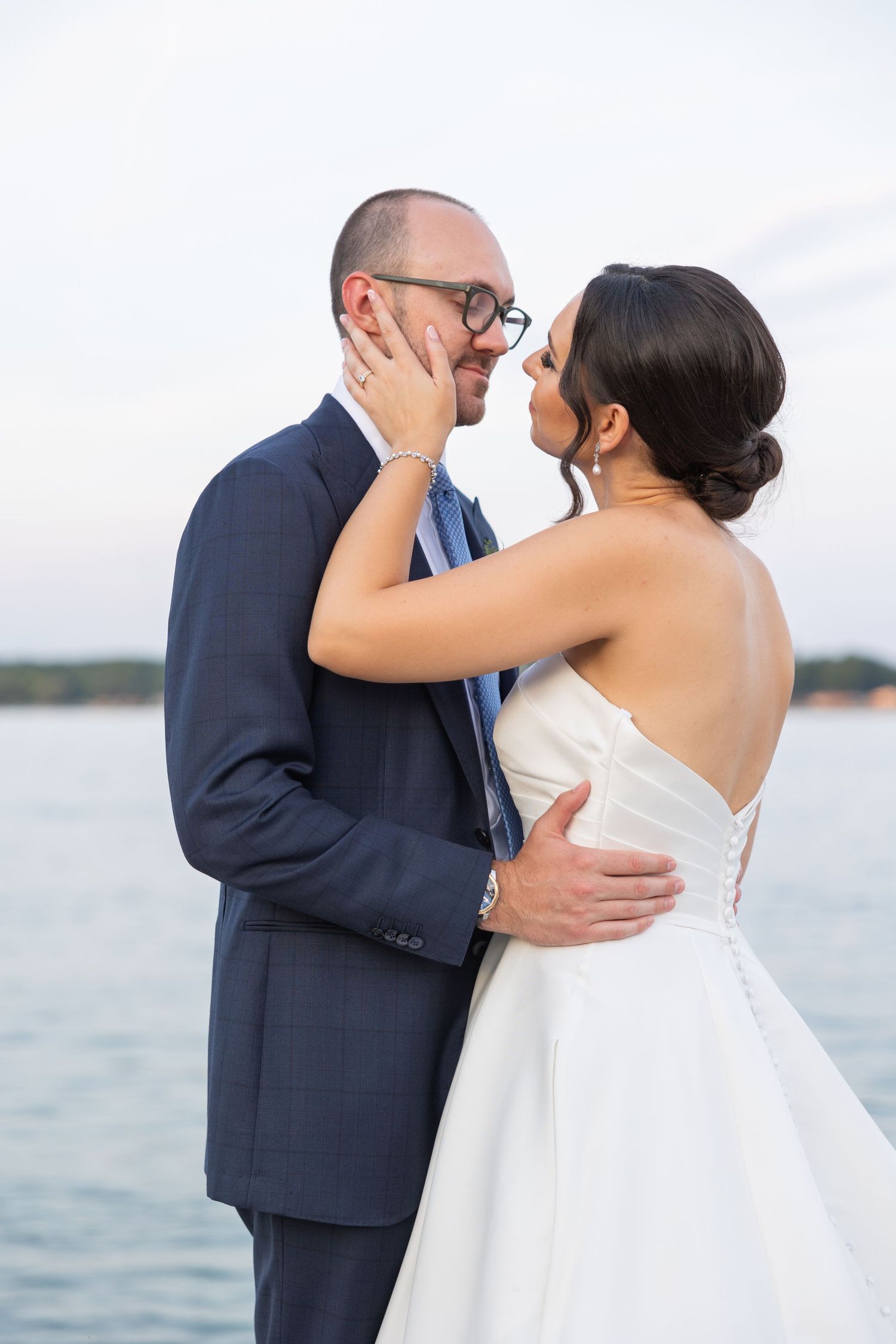 Romantic wedding couple embrace and kiss by the waterfront in an elegant navy suit and white wedding gown. Located in Metro Detroit Michigan