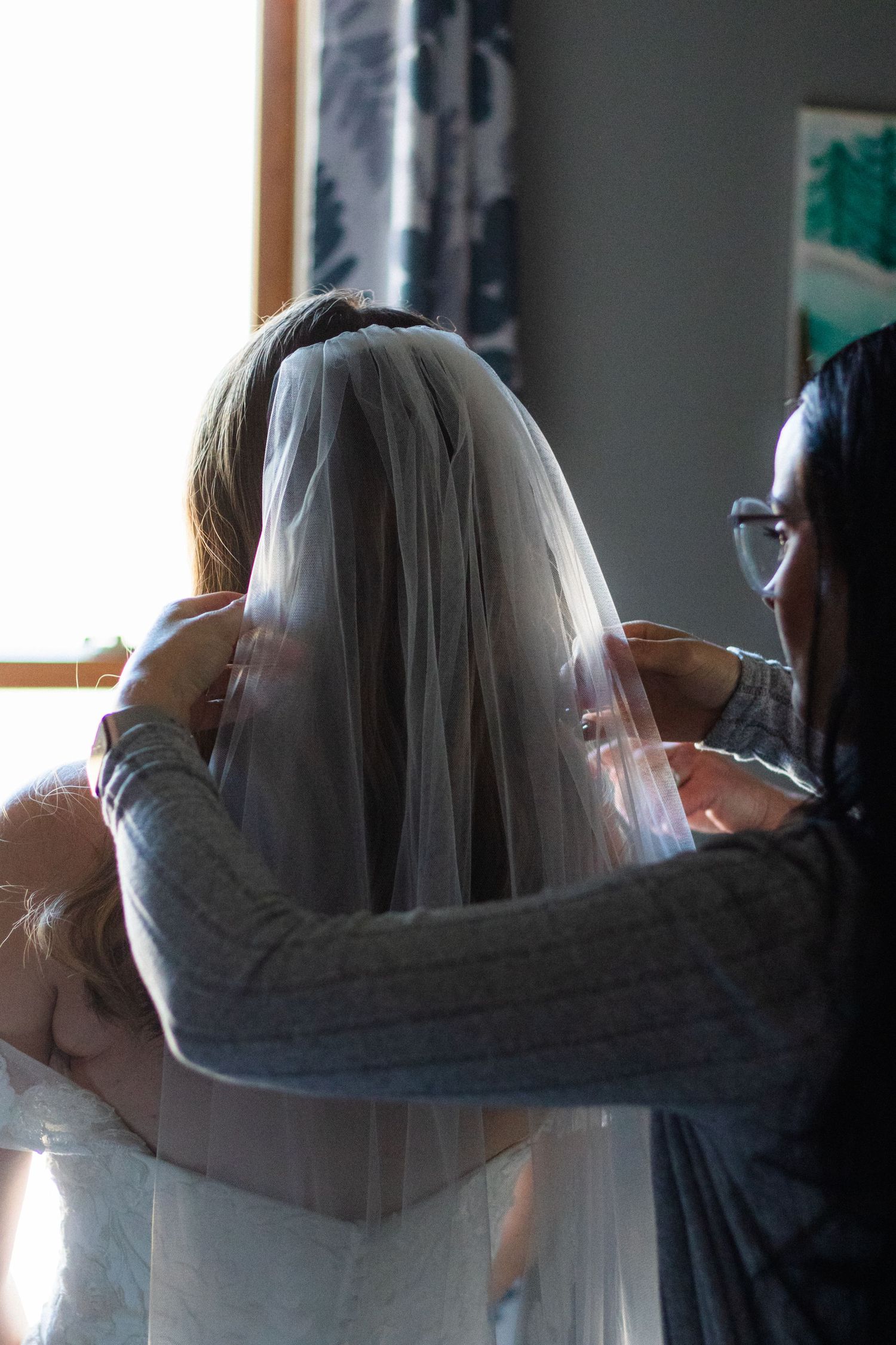 Woman in glasses helps arrange veil into brides hair as she gets ready for indoor ceremony