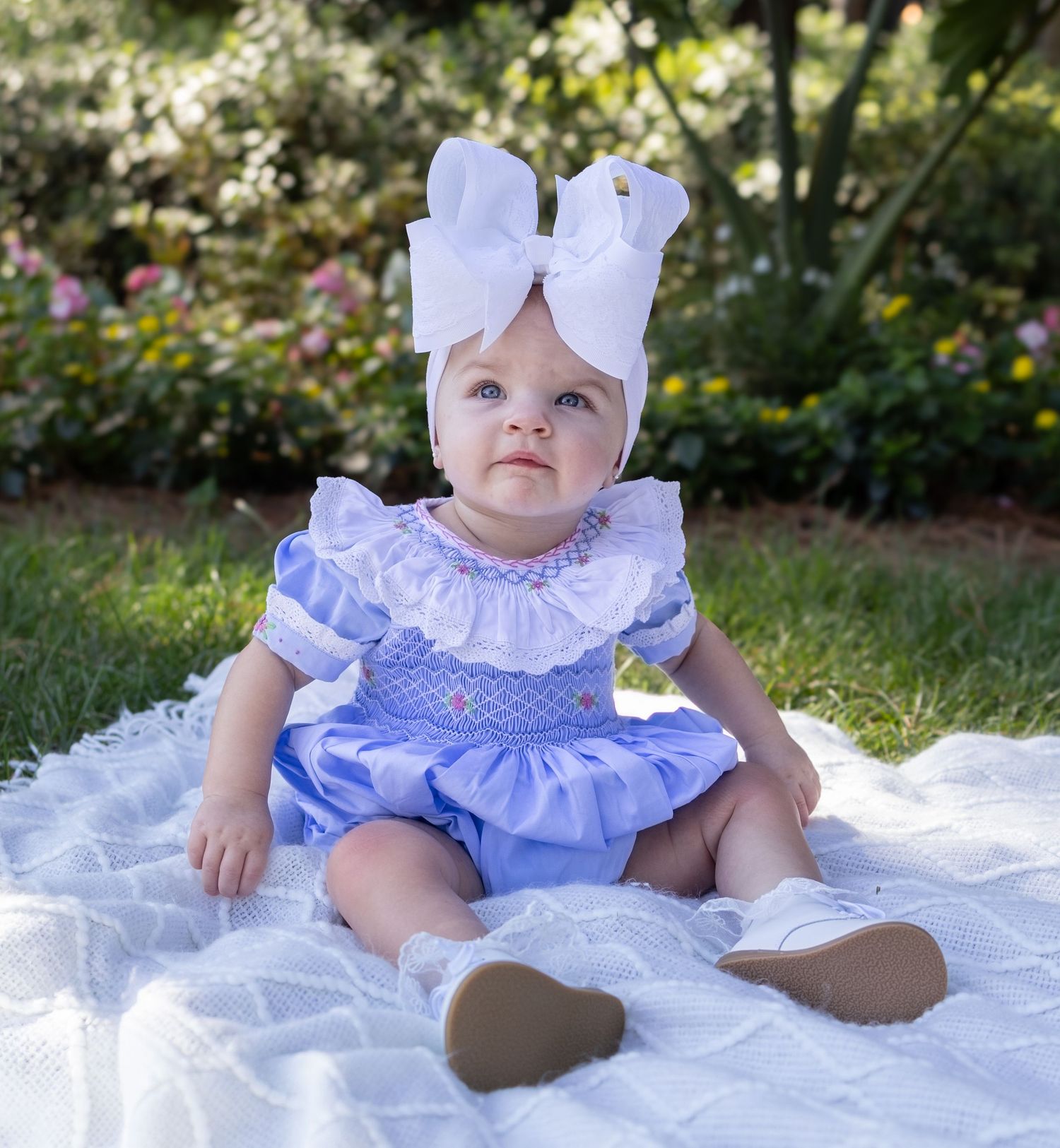 A baby in a white and blue ruffled dress sitting on a blanket outdoors.