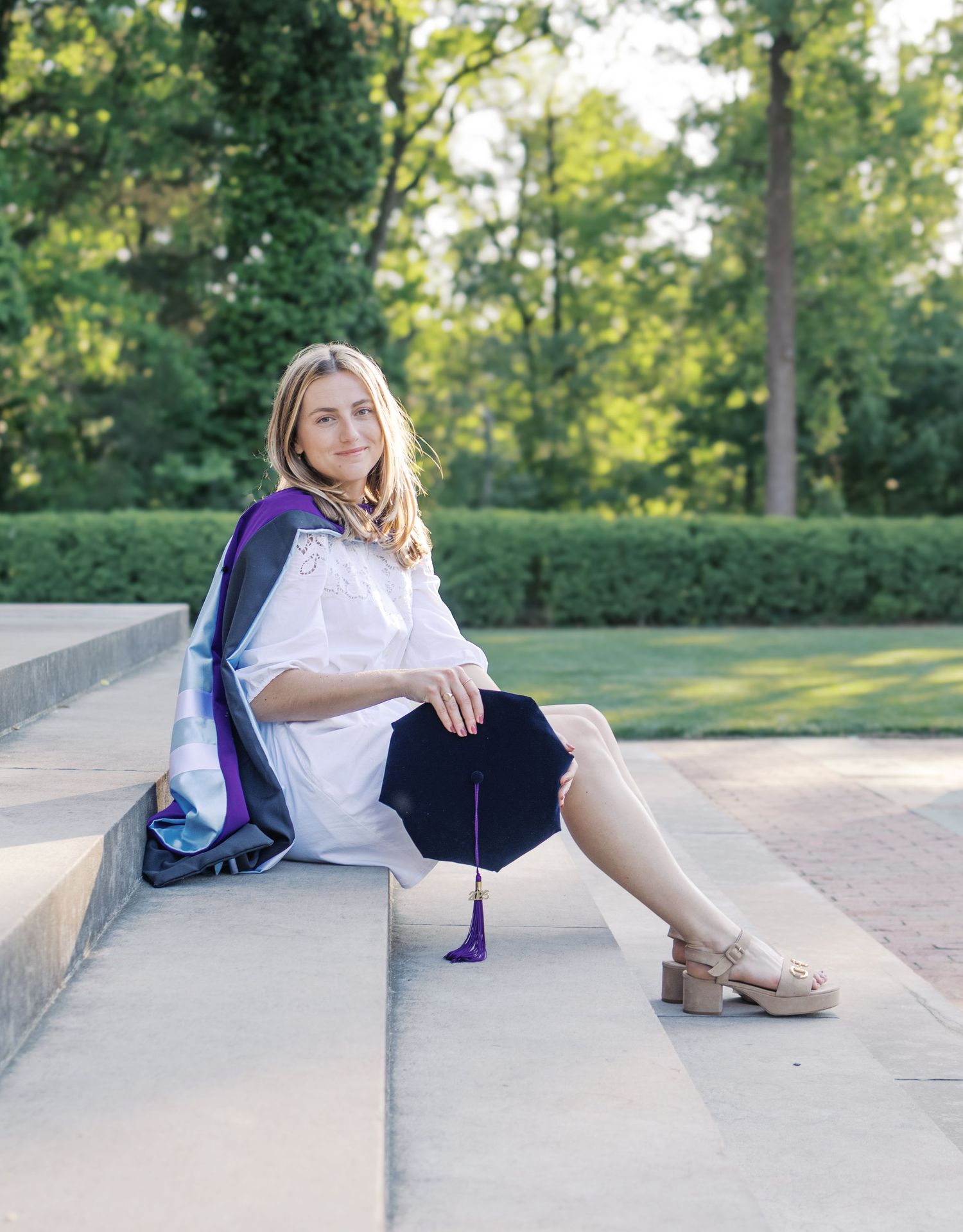 Graduate in white dress and purple stole sitting on concrete steps outdoors.