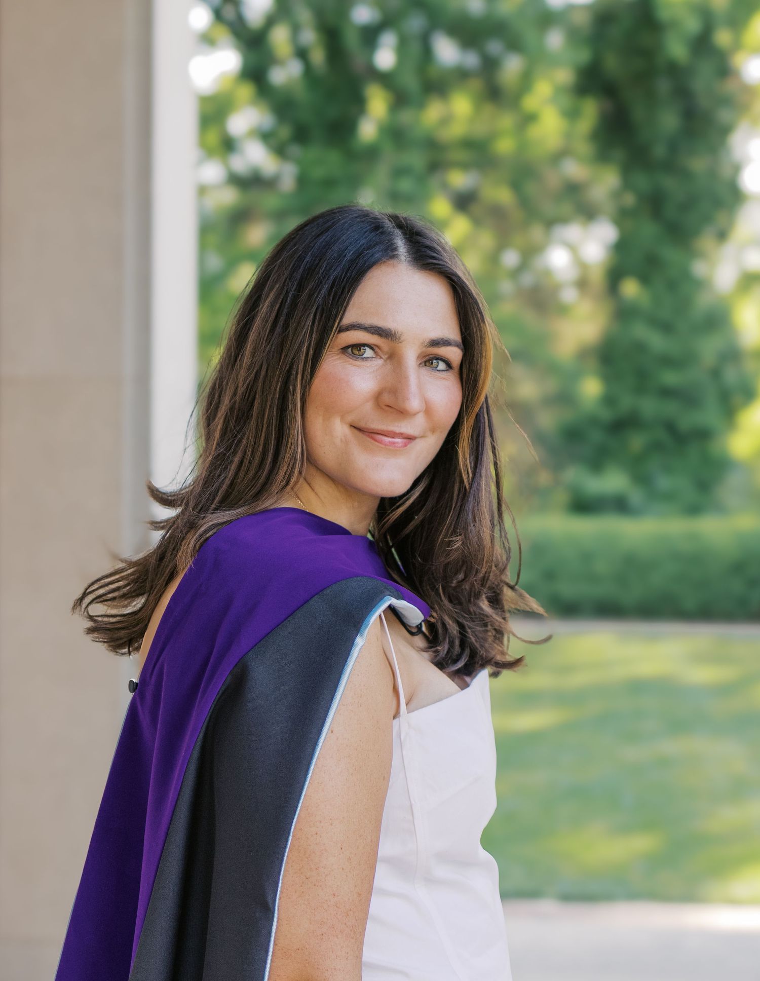 Graduate in white dress and purple stole poses outdoors on sunny day.