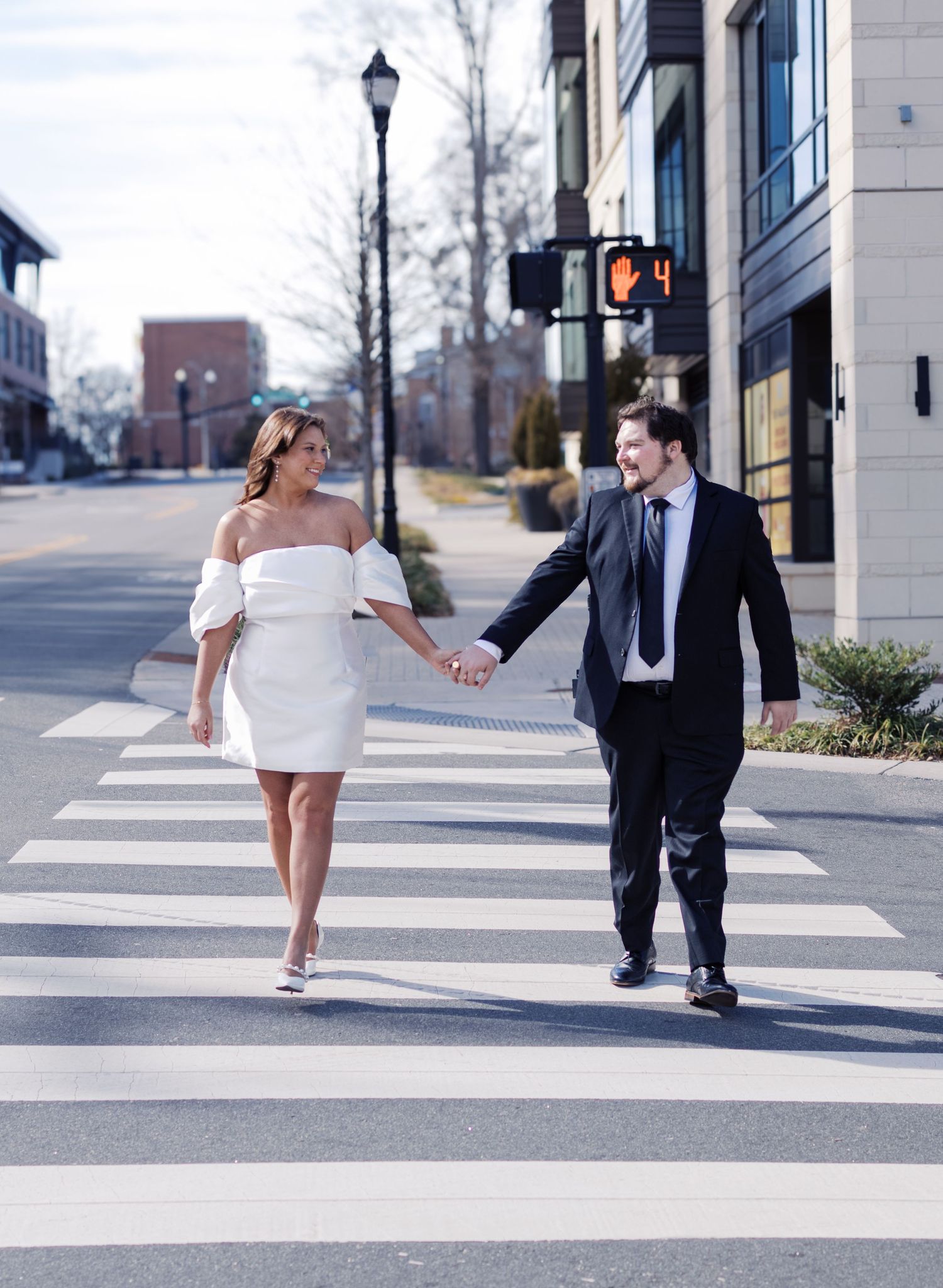 Couple in wedding attire walking playfully across a crosswalk in an urban setting.