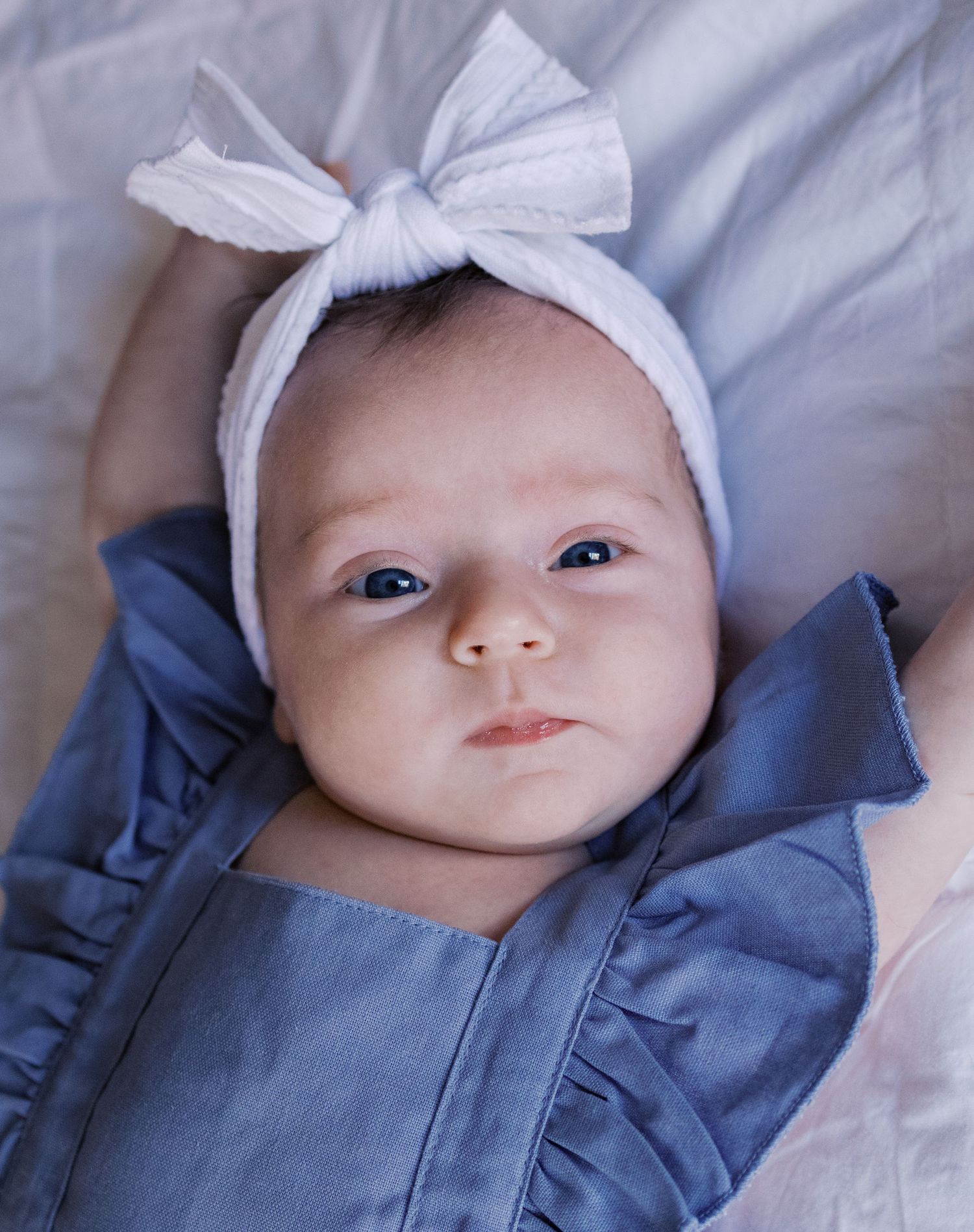 Baby wearing a white bow headband and blue ruffled outfit lying on bedding.