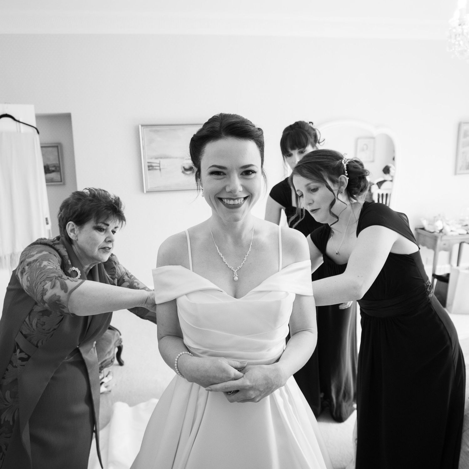 Black and white photograph showing preparations before a wedding ceremony with a bride in an elegant off-shoulder gown. Wadhurst Castle