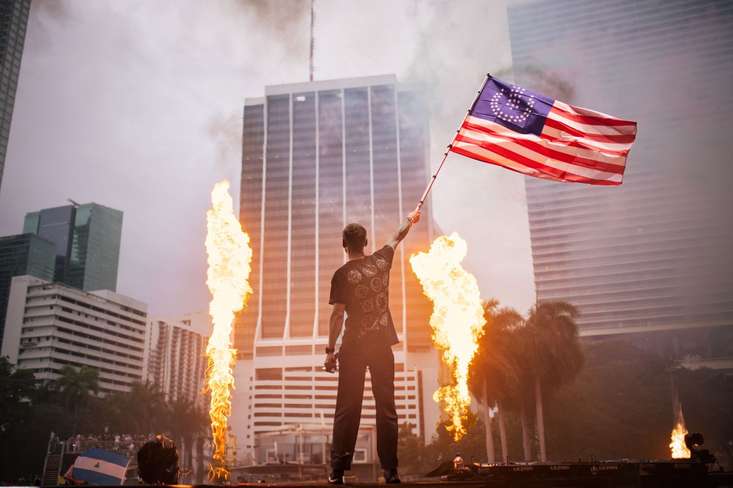 Silhouetted figure waves American flag between fire columns with city buildings in background during protest.