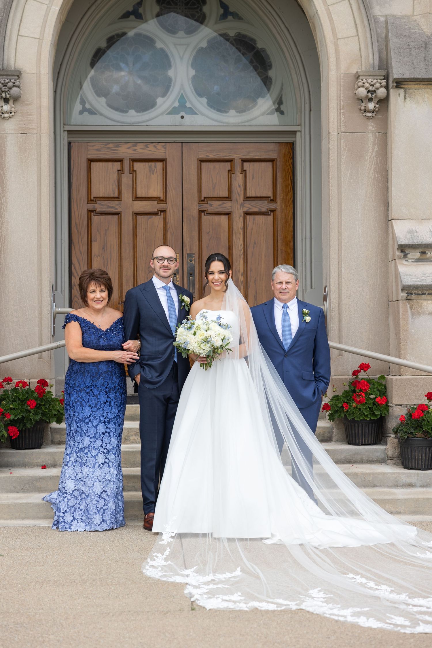 A wedding party poses in front of a grand arched church doorway with red flowers and a flowing white veil. Rachel Martinez Photography
