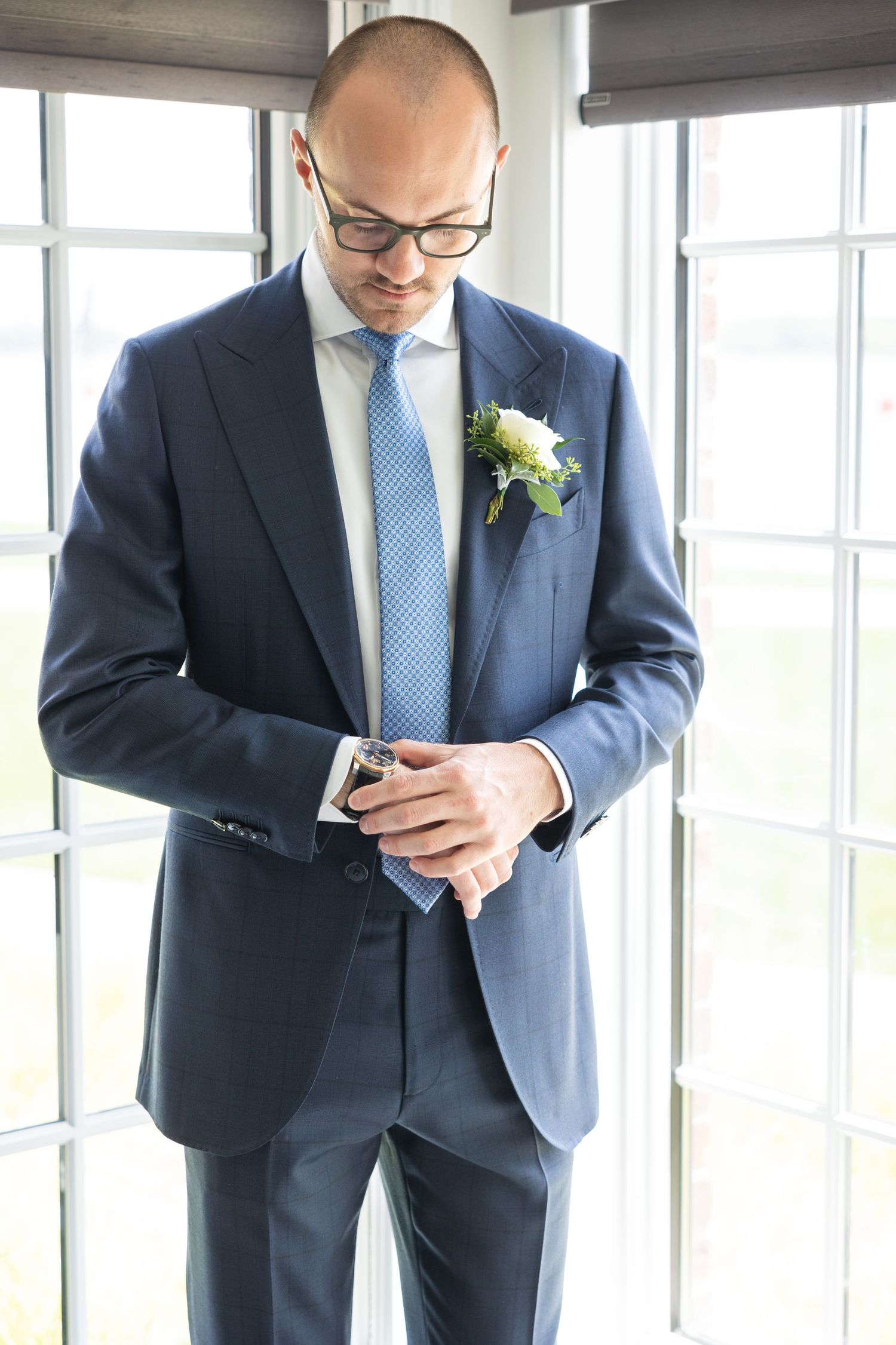 Groomsmen in navy suits and blue ties check their phones while standing near bright windows.