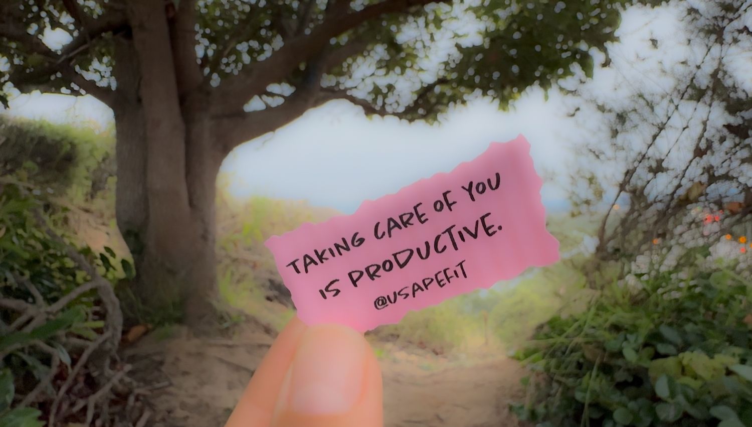 A pink sticky note with inspirational text held up against a blurred outdoor nature background.